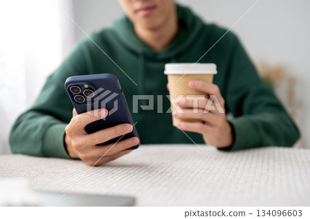 Close up of a man holding a cup of coffee and looking at phone while sitting at table in a cafe. 134096603