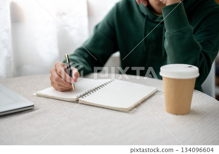 Close up of a man holding pen writing in notebook aside coffee cup while sitting at table in a cafe. 134096604