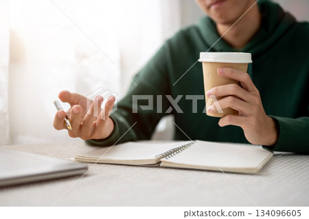Close up of a man holding coffee cup and pen over notebook aside laptop sitting at table in a cafe. 134096605