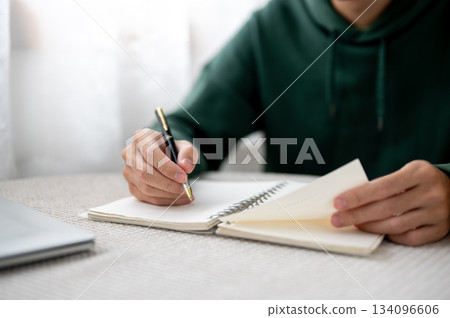 Close up of a man holding pen writing and opening a notebook aside laptop sitting at table in a cafe 134096606