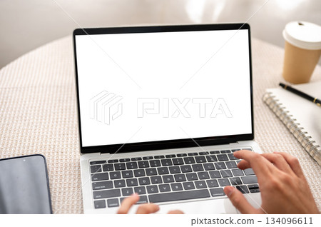Hands typing on white screen laptop aside notebook and coffee on round table in cafe or living room. 134096611