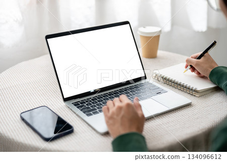 Hand typing on blank screen laptop as writing in notebook with phone on table in cafe or living room 134096612