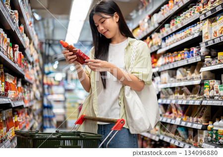 Chiang mai, Thailand - September 16th, 2025: Woman holding product over shopping cart in supermarket 134096880