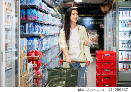 Chiang mai, Thailand - September 16th, 2025: Woman with phone pulling a shopping cart in supermarket Chiang mai, Thailand - September 16th, 2025: Woman with phone pulling a shopping cart in supermarket 134096887