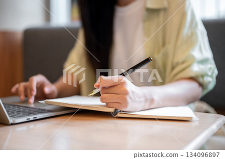 Close up woman student holding pen writing in notebook looking or typing on laptop at table in cafe. 134096897