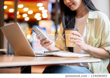 Asian woman student holding coffee looking at phone over notebook and laptop on wooden table in cafe Asian woman student holding coffee looking at phone over notebook and laptop on wooden table in cafe 134096905