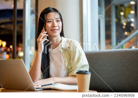 Asian woman student holding and talking on phone over notebook and laptop on wooden table in a cafe. 134096906