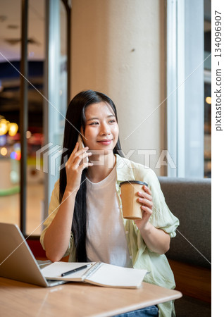 Asian woman student holding coffee talking on phone over notebook and laptop on wooden table in cafe 134096907