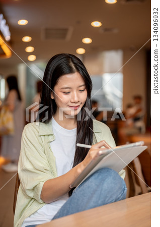 Asian woman student holding stylus writing or drawing on tablet over a knee at wooden table in cafe. Asian woman student holding stylus writing or drawing on tablet over a knee at wooden table in cafe. 134096932