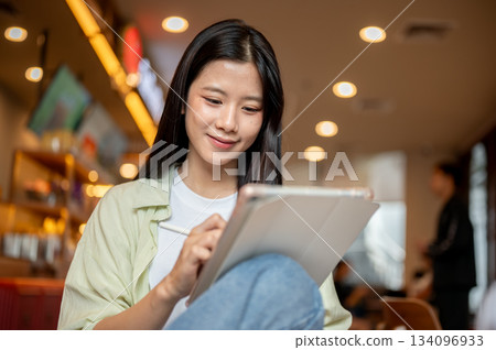 Asian woman student holding stylus writing or drawing on tablet while sitting knee up in the cafe. Asian woman student holding stylus writing or drawing on tablet while sitting knee up in the cafe. 134096933