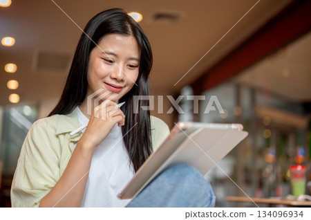Asian woman student holding stylus thinking and looking at tablet while sitting knee up in the cafe. 134096934