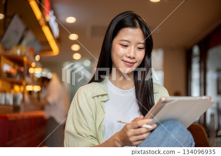 Happy asian woman student holding stylus and looking at tablet while sitting knee up in the cafe. Happy asian woman student holding stylus and looking at tablet while sitting knee up in the cafe. 134096935