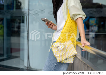 Close up woman holding phone carrying a yellow net bag standing leaning on shopping mall's railing. Close up woman holding phone carrying a yellow net bag standing leaning on shopping mall's railing. 134096945
