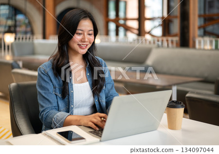 Asian woman student looking typing on laptop with phone and notebook on table in cafe or coffee shop Asian woman student looking typing on laptop with phone and notebook on table in cafe or coffee shop 134097044
