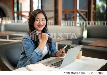 Asian woman holding phone with pen under chin sits in front of laptop aside coffee on table in cafe. Asian woman holding phone with pen under chin sits in front of laptop aside coffee on table in cafe. 134097065