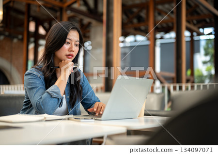 Serious asian woman holding pen under chin looking at laptop aside notebook on table sitting in cafe Serious asian woman holding pen under chin looking at laptop aside notebook on table sitting in cafe 134097071