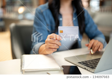 Close up woman's hand holding and showing credit card sitting with laptop and notebook at cafe table Close up woman's hand holding and showing credit card sitting with laptop and notebook at cafe table 134097083
