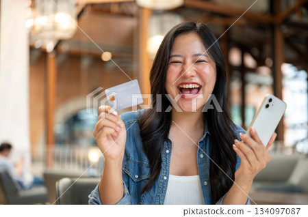 Asian woman holding credit card and phone while laughing in excited at table in cafe or coffee shop 134097087