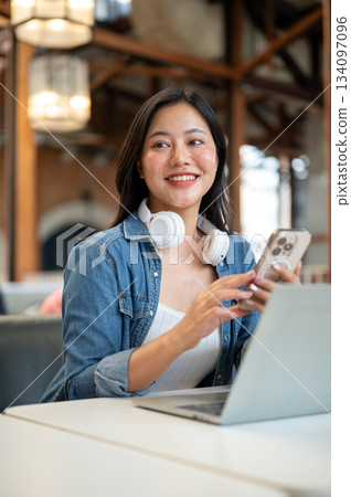 Happy asian woman with headphone looking away from phone sitting in front of laptop on table in cafe Happy asian woman with headphone looking away from phone sitting in front of laptop on table in cafe 134097096