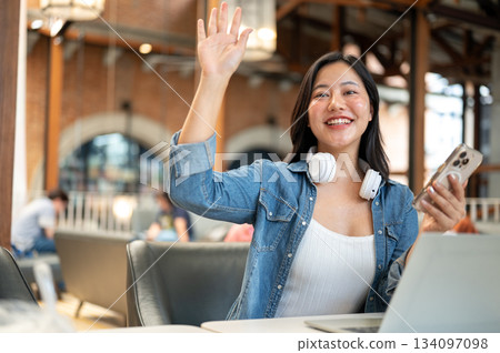 Happy asian woman holding phone and waving a hand while sitting in front of a laptop at cafe table. Happy asian woman holding phone and waving a hand while sitting in front of a laptop at cafe table. 134097098