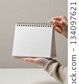 Clean tabletop calendar mockup with spiral ring. Blank paper, hand hold, soft neutral background. Perfect for brand presentation, schedule design, office stationery, planner cover. 134097621