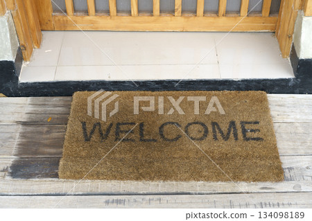 Close-up of a welcome doormat on weathered wooden planks at a doorway. The mat features the word WELCOME in black lettering. The entrance has a tiled threshold and a wooden door frame. Close-up of a welcome doormat on weathered wooden planks at a doorway. The mat features the word WELCOME in black lettering. The entrance has a tiled threshold and a wooden door frame. 134098189