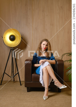 Young woman sitting in a chair wearing blue dress in a simple interior with a lamp Young woman sitting in a chair wearing blue dress in a simple interior with a lamp 134098281