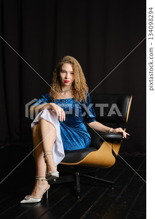 Model poses in a chair wearing a blue dress with a cigarette in hand during a studio session Model poses in a chair wearing a blue dress with a cigarette in hand during a studio session 134098294