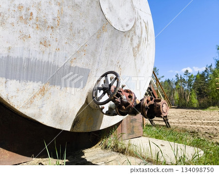 Old rusty industrial storage tank with large valve. Close-up of a weathered white industrial storage tank with heavy rust spots, featuring a large manual valve wheel in an outdoor setting. Old rusty industrial storage tank with large valve. Close-up of a weathered white industrial storage tank with heavy rust spots, featuring a large manual valve wheel in an outdoor setting. 134098750