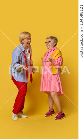 Two senior women laughing together on yellow background. 134099121