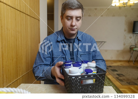 Man taking dietary supplements at home, pouring pills from a bottle and drinking with water. Concept of daily vitamins, health care and wellness routine. Man taking dietary supplements at home, pouring pills from a bottle and drinking with water. Concept of daily vitamins, health care and wellness routine. 134099328