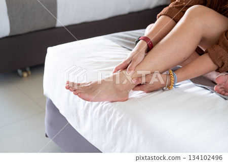 Asian woman practicing gentle ankle massage on bed, highlighting comfort and home wellness 134102496