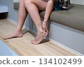 Asian woman sitting beside her bed massaging her ankle for pain relief, calm and quiet home moment 134102499