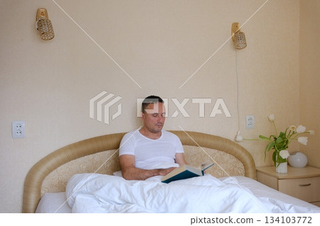 Man reading book in bedroom feeling serene and relaxed mood at night closeup. Engaged male reader absorbed in literature at lamp light. Calm guy enjoy hobby lying bed in tranquil home atmosphere. 134103772