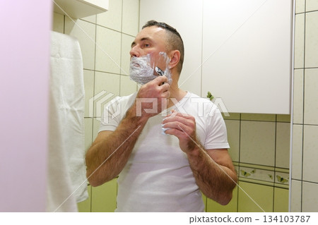 A man in the bathroom in front of a mirror applies shaving cream to his face. A man in the bathroom in front of a mirror applies shaving cream to his face. 134103787