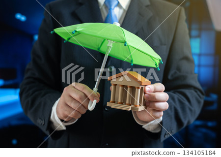 Businessman holds a green umbrella over a bank or government building. Protection of investments and financial well-being. Investment security, bank insurance, safe savings, and risk management 134105184
