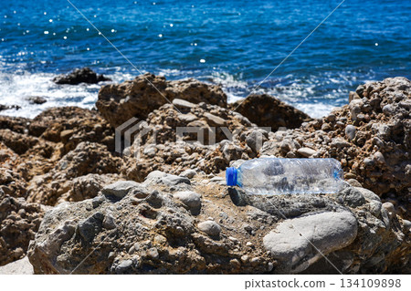 An Oversized Plastic Bottle Discarded Amidst Rugged Rocks by the Shore, Highlighting Ocean Pollution and Environmental Concerns 134109898