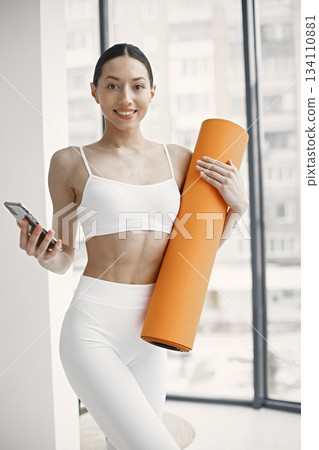 Slim and beautiful woman wearing white leggins and top and holding rolling yoga mat. Young girl with fit body posing in studio with big windows. Sport, fitness, diet, nutrition and health care concept 134110881