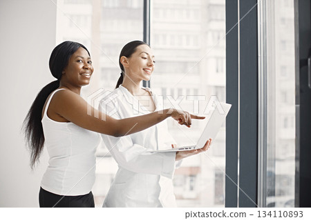 Young female caucasian doctor standing in her office at clinic near big windows with her female black patient. Woman wearing white medical coat. Women using a laptop. Young female caucasian doctor standing in her office at clinic near big windows with her female black patient. Woman wearing white medical coat. Women using a laptop. 134110893