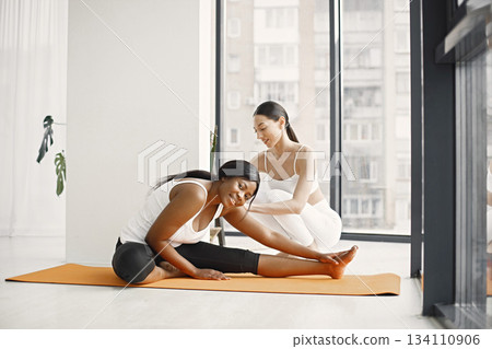 Two women working out on yoga mat in bright studio with big windows. Caucasian woman wearing white leggins and top and black woman white t-shirt and black leggins. Woman and her coach in gym. 134110906