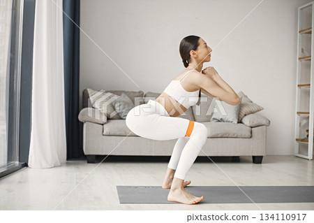 Attractive young woman doing excercises with fitness rubber band at home. Brunette woman wearing white leggins and top. Self-isolation is beneficial, entertainment and education on the Internet. Attractive young woman doing excercises with fitness rubber band at home. Brunette woman wearing white leggins and top. Self-isolation is beneficial, entertainment and education on the Internet. 134110917