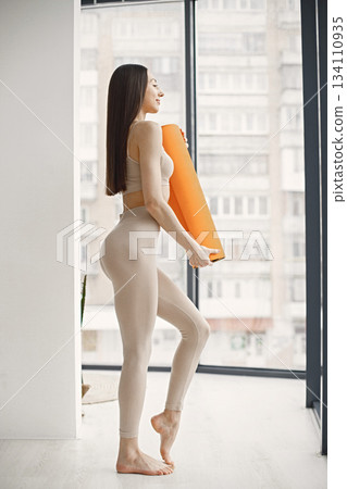 Slim and beautiful woman wearing beige leggins and top and holding rolling yoga mat. Young girl with fit body posing in studio with big windows. Sport, fitness, diet, nutrition and health care concept Slim and beautiful woman wearing beige leggins and top and holding rolling yoga mat. Young girl with fit body posing in studio with big windows. Sport, fitness, diet, nutrition and health care concept 134110935
