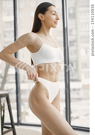 Slim and beautiful woman in white underwear. Young girl with fit body posing in underwear in studio with big windows. Sport, fitness, diet, nutrition and health care concept. Slim and beautiful woman in white underwear. Young girl with fit body posing in underwear in studio with big windows. Sport, fitness, diet, nutrition and health care concept. 134110938