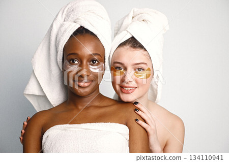Two young women in white bathrobes standing isolated on white. Girls wearing white towels on their hair. Girls have a eye patches on their faces. Two young women in white bathrobes standing isolated on white. Girls wearing white towels on their hair. Girls have a eye patches on their faces. 134110941