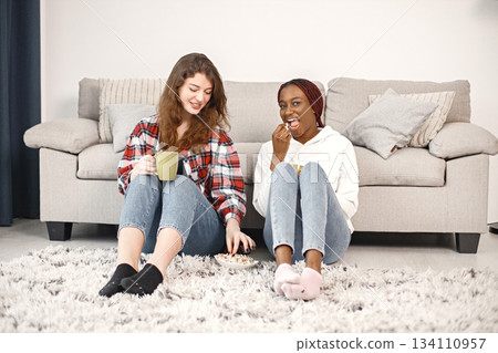Two young woman sitting on a floor near sofa and talking. Black girl wearing white hoodie, caucasian girl wearing plaid shirt. Girls holding a green cups with tea. Two young woman sitting on a floor near sofa and talking. Black girl wearing white hoodie, caucasian girl wearing plaid shirt. Girls holding a green cups with tea. 134110957