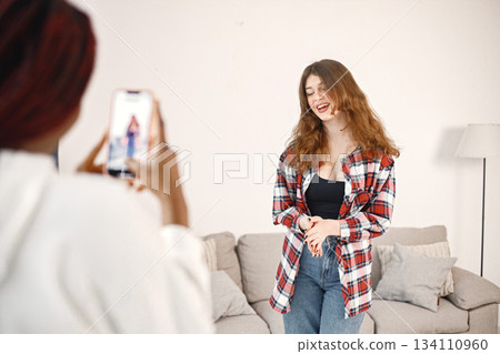 Focus on a caucasian girl who posing. Two young woman standing near coach at home. Black girl wearing white hoodie, caucasian girl wearing plaid shirt. Focus on a caucasian girl who posing. Two young woman standing near coach at home. Black girl wearing white hoodie, caucasian girl wearing plaid shirt. 134110960