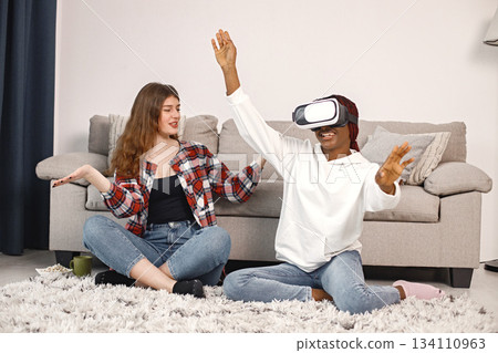 Two young women sitting on a floor near bed wearing virtual reality glasses. Black girl wearing white hoodie and caucasian girl wearing plaid shirt. Girls playing games together. Two young women sitting on a floor near bed wearing virtual reality glasses. Black girl wearing white hoodie and caucasian girl wearing plaid shirt. Girls playing games together. 134110963
