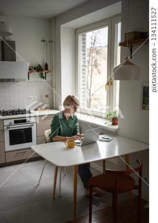 Relaxed middle aged business woman comfortably working typing on laptop online in kitchen. 134111527