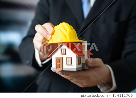 A man puts a yellow construction helmet on a house. House renovation. Building reconstruction. Structural repair modernization. Construction industry updates, property maintenance, home improvement. 134111706