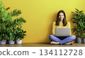 Young Woman with Laptop Sitting on Floor Surrounded by Green Plants 134112728